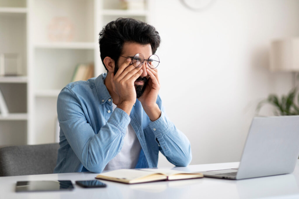 Glaucoma Concept. Young Indian Man Massaging Tired Eyes After Working On Laptop Computer, Millennial Eastern Male Suffering From Eyestrain And Fatigue, Sitting At Workplace In Home Office